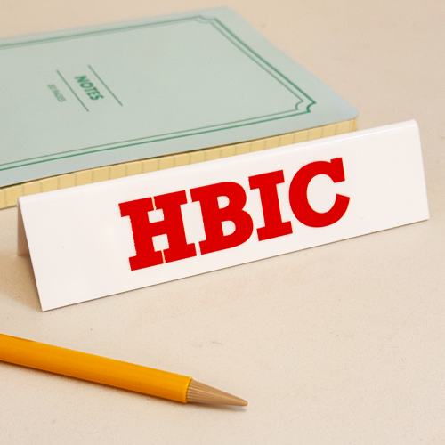 White desk sign with the letters H B I C in vibrant red, for Head Bitch In Charge.