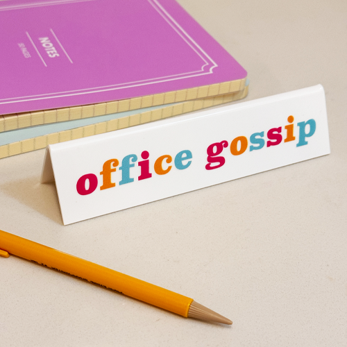 Tent-style desk sign with the words Office Gossip written on it in different colored letters.