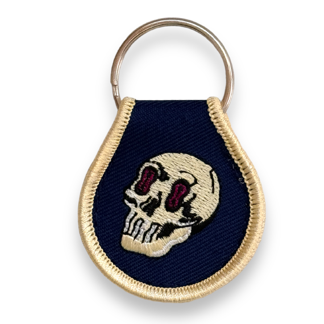 Dark blue droplet-shaped keychain embroidered with gold border and gold skull.